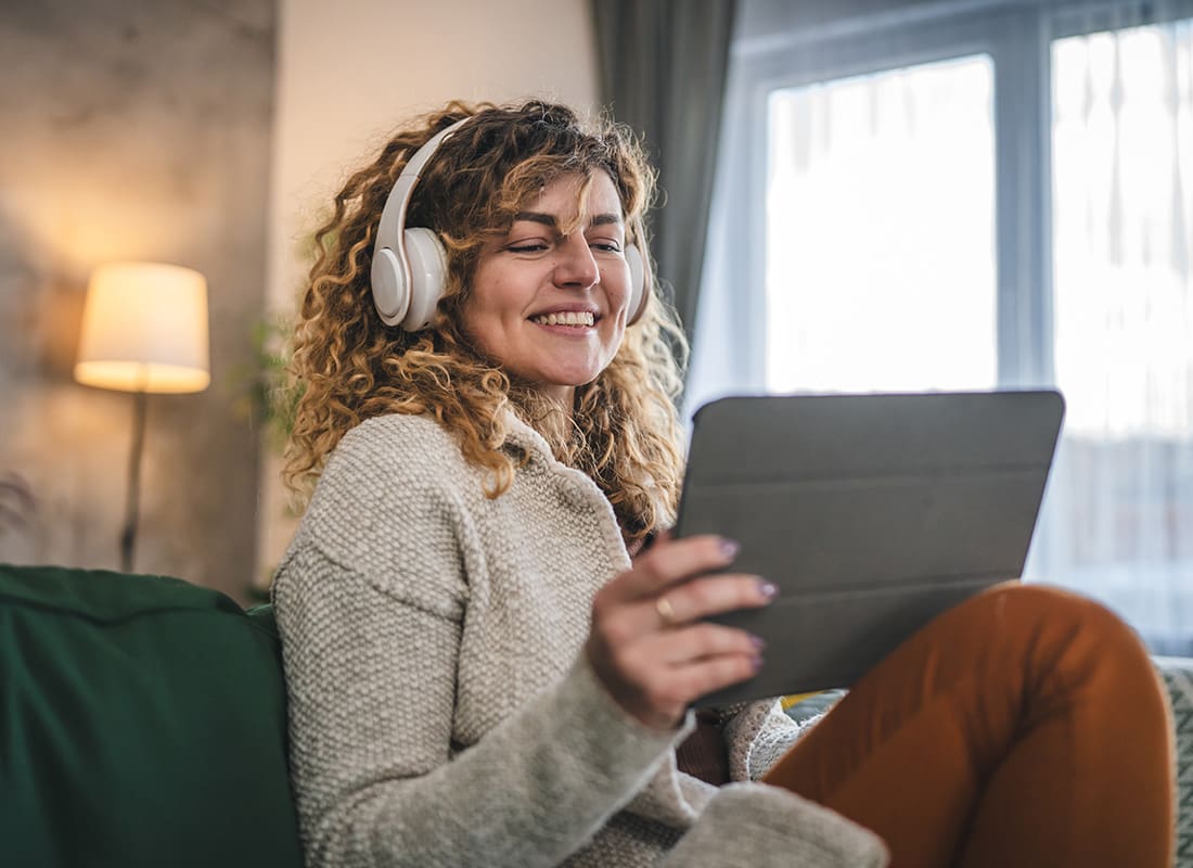 Video Library - Portrait of a Cheerful Middle Aged Woman Wearing Headphones Sitting on the Sofa Watching Videos on a Tablet